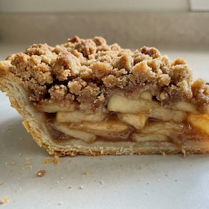 Può includere: Una fetta di torta di mele con crumble su un piatto chiaro. La torta presenta strati di mele a fette, una crosta friabile e una copertura sbriciolata marrone dorato. La crosta è dorata e le mele sono di colore giallo chiaro.