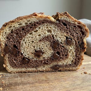 May include: A cross-section of chocolate babka bread, showcasing a swirl of chocolate and dough. The bread has a golden-brown crust and visible chocolate chips throughout. The loaf sits on a wooden cutting board.