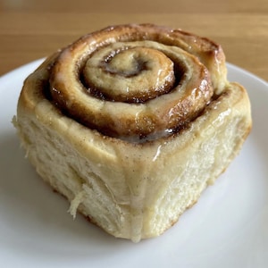 Könnte beinhalten: Ein frisch gebackenes Zimtbrötchen mit glänzendem Zuckerguss, serviert auf einem weißen Teller. Das spiralförmige Design des Brötchens ist sichtbar, mit einer goldbraunen Außenseite und einem weichen, fluffigen Inneren. Der Guss hat winzige Vanille-Bohnen-Punkte.