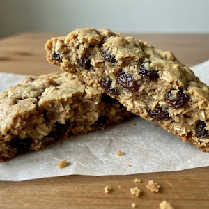 Puede incluir: Primer plano de dos mitades de una galleta grande de avena y pasas. La galleta es de color marrón claro con avena visible y pasas oscuras. Se encuentra sobre papel de hornear.