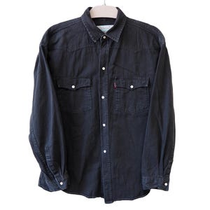 Pode incluir: Camisa de ganga escura com dois bolsos no peito e botões brancos. A camisa de manga comprida tem uma gola clássica e está pendurada num cabide. Uma pequena etiqueta vermelha é visível no bolso esquerdo.