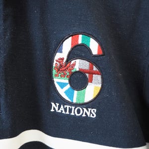 May include: Navy blue long-sleeve shirt with a white and black striped design. The shirt features an embroidered emblem of the number "6" with flags and the word "NATIONS" in white.