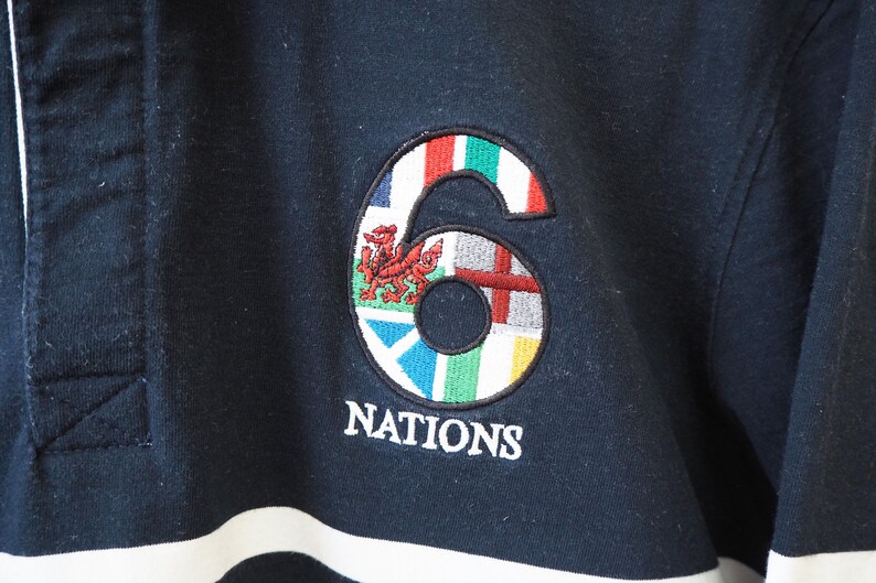 May include: Navy blue long-sleeve shirt with a white and black striped design. The shirt features an embroidered emblem of the number "6" with flags and the word "NATIONS" in white.