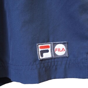 May include: Close-up of navy blue fabric with a stitched-on Fila logo patch. The patch features the brand name in a white circle and the Fila logo in red, white, and black. The fabric appears to be part of a garment.