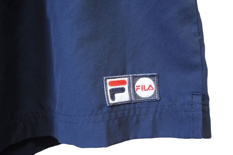 May include: Close-up of navy blue fabric with a stitched-on Fila logo patch. The patch features the brand name in a white circle and the Fila logo in red, white, and black. The fabric appears to be part of a garment.