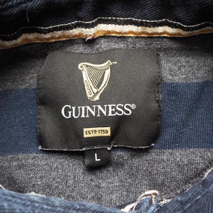 Puede incluir: Primer plano de una camisa a rayas azul oscuro y gris con el logotipo de Guinness en una etiqueta negra. La etiqueta incluye un s&iacute;mbolo de arpa y el texto "ESTD 1759". La camisa es talla L.
