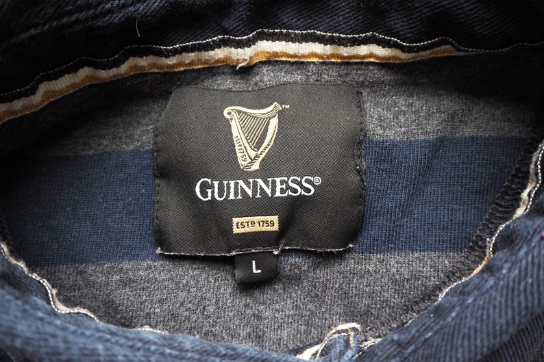 Puede incluir: Primer plano de una camisa a rayas azul oscuro y gris con el logotipo de Guinness en una etiqueta negra. La etiqueta incluye un s&iacute;mbolo de arpa y el texto "ESTD 1759". La camisa es talla L.