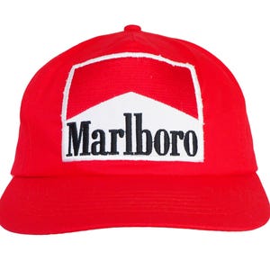 Può includere: Un cappellino da baseball rosso brillante con una toppa rettangolare sulla parte anteriore. La toppa presenta uno sfondo bianco con uno chevron rosso nella parte superiore e la parola Marlboro stampata in grassetto nero al centro.