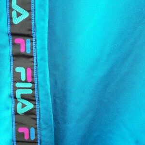 May include: Close-up of a turquoise fabric with a vertical black stripe featuring the brand name "FILA" in teal, pink, and white. The stripe is bordered by blue stitching, and the background is a solid turquoise color.