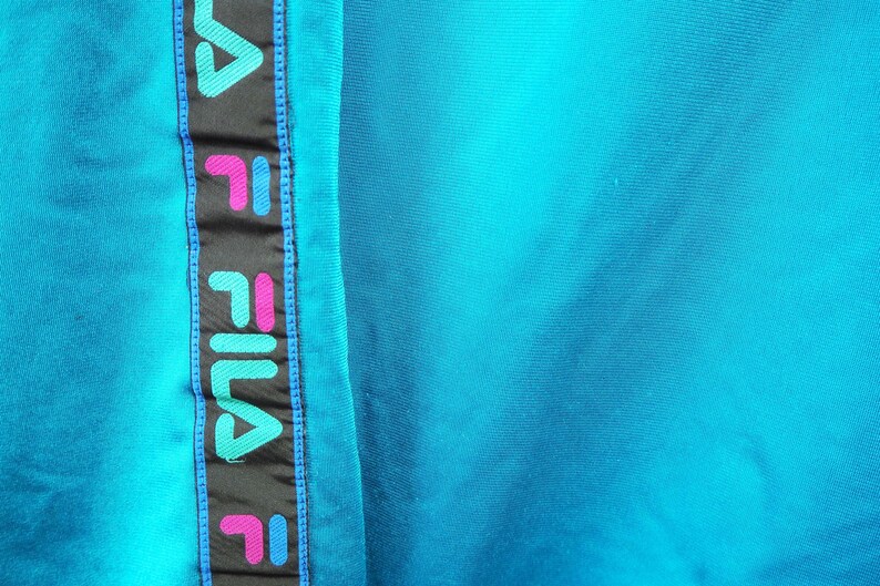 May include: Close-up of a turquoise fabric with a vertical black stripe featuring the brand name "FILA" in teal, pink, and white. The stripe is bordered by blue stitching, and the background is a solid turquoise color.