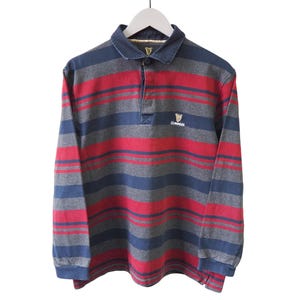 Puede incluir: Una camiseta de rugby de manga larga con rayas horizontales en rojo, azul y gris. La camiseta tiene cuello y un peque&ntilde;o logotipo de Guinness en el pecho. La camiseta est&aacute; colgada de una percha blanca. Medidas en cent&iacute;metros.