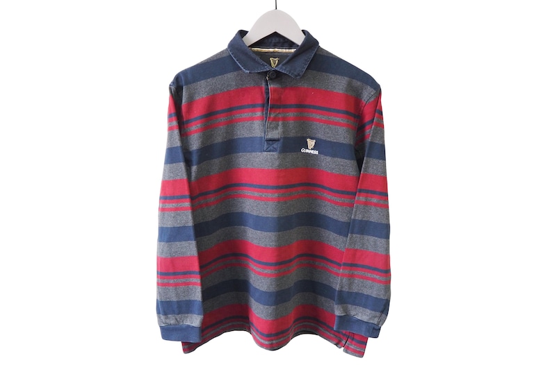 Puede incluir: Una camiseta de rugby de manga larga con rayas horizontales en rojo, azul y gris. La camiseta tiene cuello y un peque&ntilde;o logotipo de Guinness en el pecho. La camiseta est&aacute; colgada de una percha blanca. Medidas en cent&iacute;metros.