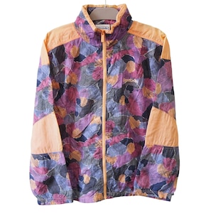 Może przedstawiać: Wiatrówka Reebok w stylu vintage. Kurtka ma kolorowy kwiatowy wzór w odcieniach fioletu, niebieskiego i pomarańczowego. Kołnierz i panele na ramionach są w jednolitym brzoskwiniowym kolorze. Logo Reebok jest widoczne na piersi.