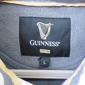 May include: Close-up of a gray fleece jacket with a black label featuring the Guinness logo, a harp, and the text "GUINNESS" and "ESTD 1759". The jacket is size L.