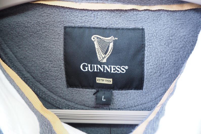 May include: Close-up of a gray fleece jacket with a black label featuring the Guinness logo, a harp, and the text "GUINNESS" and "ESTD 1759". The jacket is size L.