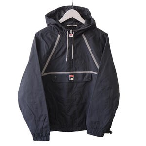 Op de afbeelding: Zwarte anorakjas met capuchon, hangend aan een witte hanger. De jas heeft een voorzak, een halve ritssluiting en reflecterende zilveren strepen. Het Fila-logo is zichtbaar op de zak. Een casual bovenkledingstuk.