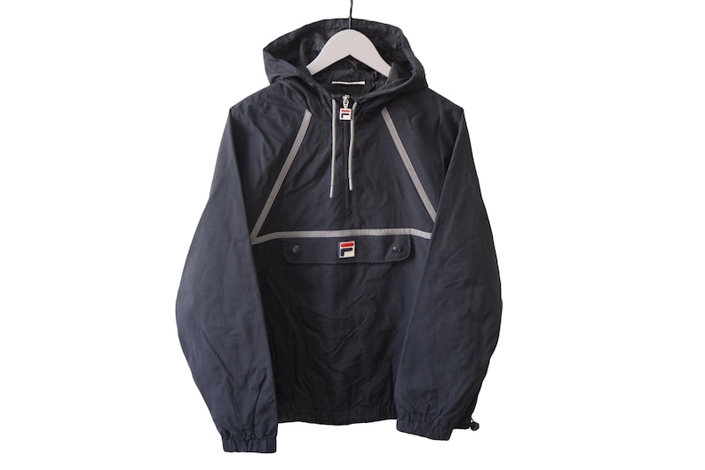 Op de afbeelding: Zwarte anorakjas met capuchon, hangend aan een witte hanger. De jas heeft een voorzak, een halve ritssluiting en reflecterende zilveren strepen. Het Fila-logo is zichtbaar op de zak. Een casual bovenkledingstuk.