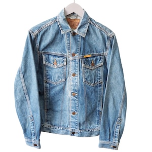 Puede incluir: Chaqueta vaquera azul claro con botones, dos bolsillos en el pecho y dos bolsillos laterales. La chaqueta tiene un cuello cl&aacute;sico y est&aacute; colgada de una percha blanca. La chaqueta tiene una peque&ntilde;a etiqueta amarilla en el bolsillo del pecho.