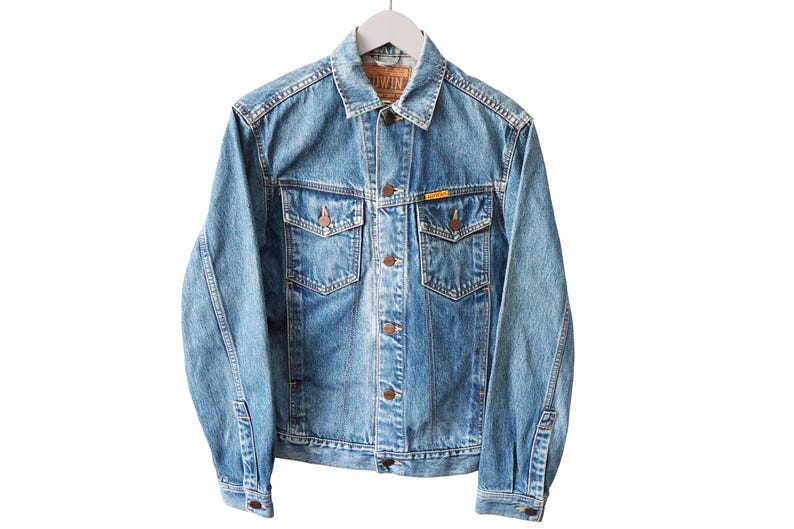 Puede incluir: Chaqueta vaquera azul claro con botones, dos bolsillos en el pecho y dos bolsillos laterales. La chaqueta tiene un cuello cl&aacute;sico y est&aacute; colgada de una percha blanca. La chaqueta tiene una peque&ntilde;a etiqueta amarilla en el bolsillo del pecho.
