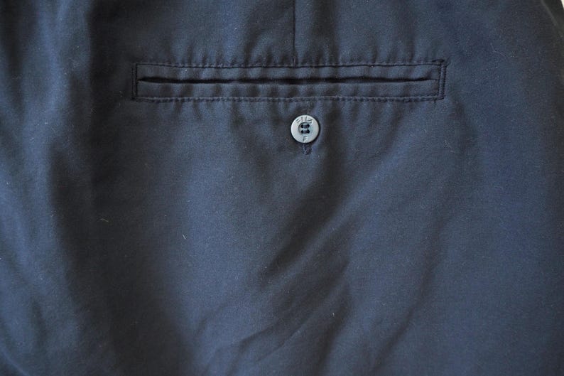 May include: Close-up of navy blue fabric with a pocket and button detail. The pocket is rectangular and stitched into the fabric. A small, round button is centered below the pocket.