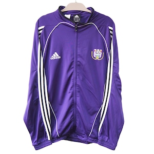 Puede incluir: Chaqueta deportiva Adidas morada con rayas blancas en las mangas y el logo blanco de Adidas en el pecho. Cuenta con cremallera completa y escudo del equipo. La chaqueta está colgada en una percha.