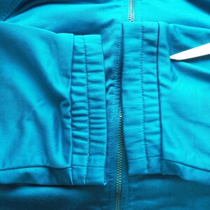 May include: Close-up of a teal-colored athletic jacket. The jacket features a full-length zipper and detailed stitching along the seams. A pair of scissors is placed on the jacket.