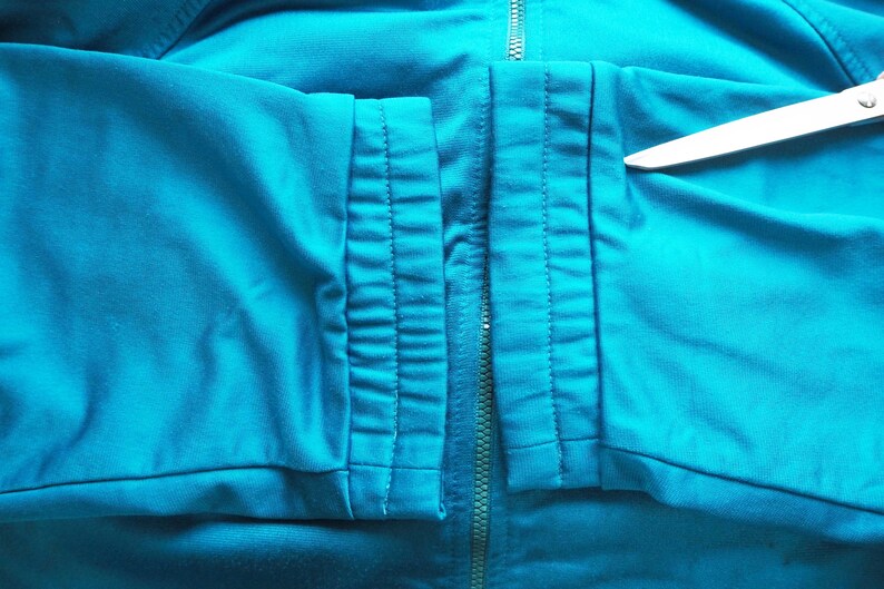 May include: Close-up of a teal-colored athletic jacket. The jacket features a full-length zipper and detailed stitching along the seams. A pair of scissors is placed on the jacket.