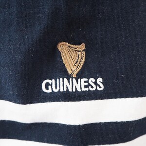 May include: Close-up of a navy blue and white striped garment. The garment features the Guinness logo embroidered in gold and the word "GUINNESS" in white. The fabric appears to be a soft knit.