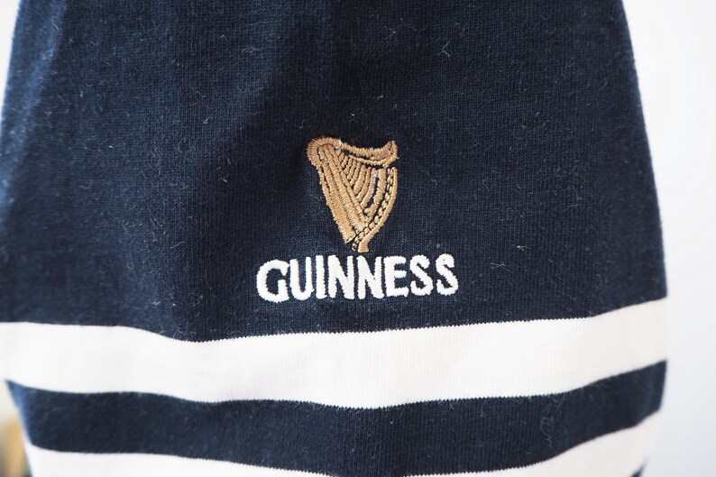 May include: Close-up of a navy blue and white striped garment. The garment features the Guinness logo embroidered in gold and the word "GUINNESS" in white. The fabric appears to be a soft knit.