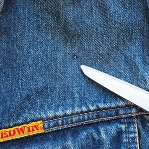 Puede incluir: Primer plano de una chaqueta vaquera azul con una etiqueta amarilla y roja "EDWIN". Una cuchilla blanca est&aacute; colocada diagonalmente sobre la mezclilla. La chaqueta tiene costuras visibles y un peque&ntilde;o agujero.
