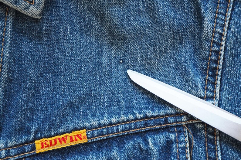Puede incluir: Primer plano de una chaqueta vaquera azul con una etiqueta amarilla y roja "EDWIN". Una cuchilla blanca est&aacute; colocada diagonalmente sobre la mezclilla. La chaqueta tiene costuras visibles y un peque&ntilde;o agujero.