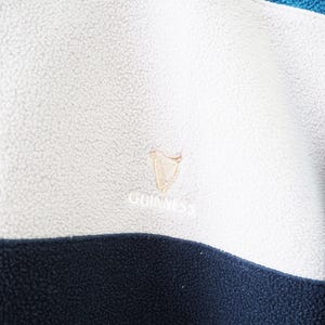 以下が含まれることがあります： 白、ネイビーブルー、ティール色のフリースプルオーバーのクローズアップ。ギネスのロゴが金色の糸で刺繍されています。フリース素材は質感があり、柔らかく見えます。暖かくカジュアルな衣服。