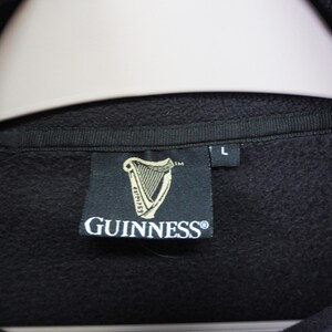 May include: Black sweatshirt with a tag featuring the Guinness logo, a golden harp, and the Guinness brand name. The tag also has the size "L" printed on it. The sweatshirt is hanging on a white hanger.