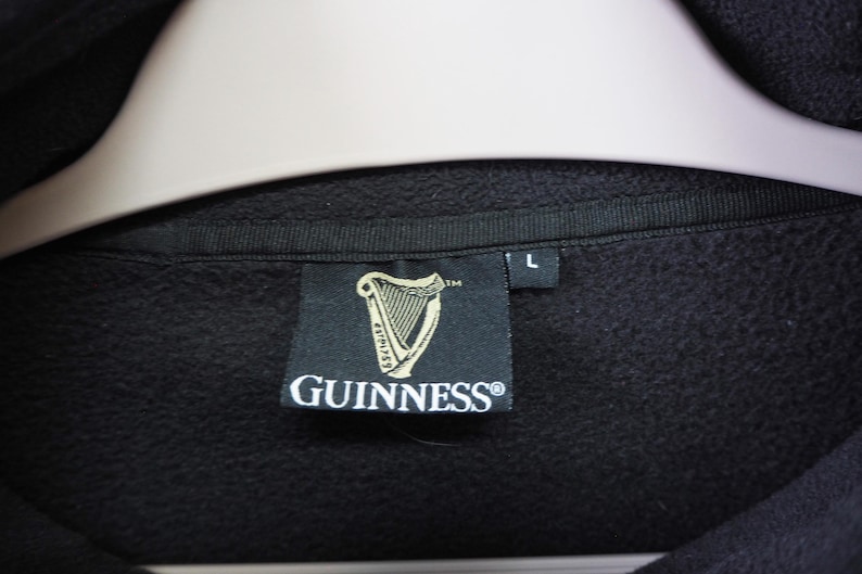 May include: Black sweatshirt with a tag featuring the Guinness logo, a golden harp, and the Guinness brand name. The tag also has the size "L" printed on it. The sweatshirt is hanging on a white hanger.