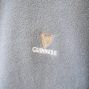 May include: Close-up of a gray fleece garment with the Guinness logo embroidered on the chest. The logo features a gold harp and the word "Guinness" in white lettering. The fleece has a textured surface.
