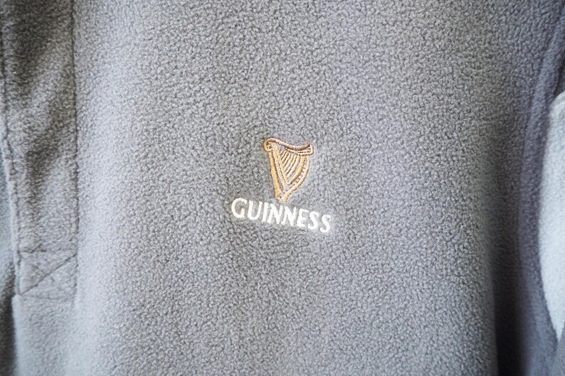 May include: Close-up of a gray fleece garment with the Guinness logo embroidered on the chest. The logo features a gold harp and the word "Guinness" in white lettering. The fleece has a textured surface.