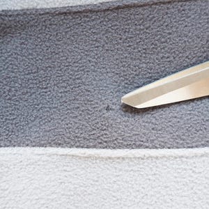 May include: Close-up of a gray, white, and gray striped fleece fabric. The fabric has a soft, textured appearance. A pair of silver scissors is positioned diagonally across the fabric, with the blades in the upper right corner.
