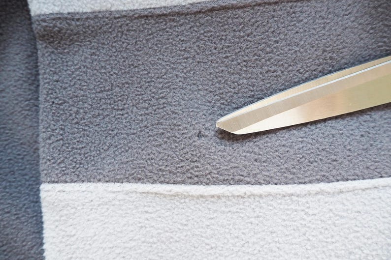 May include: Close-up of a gray, white, and gray striped fleece fabric. The fabric has a soft, textured appearance. A pair of silver scissors is positioned diagonally across the fabric, with the blades in the upper right corner.
