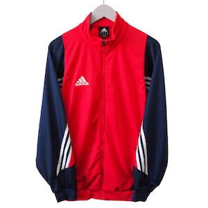 Peut inclure: Veste de survêtement Adidas rouge et bleu marine avec fermeture éclair intégrale. La veste présente des rayures blanches sur les manches et le logo Adidas sur la poitrine. Accrochée à un cintre blanc.