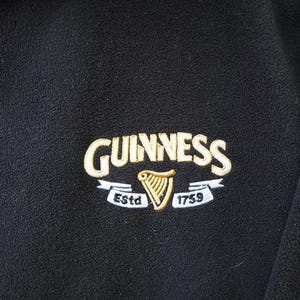 May include: Black fleece jacket with the Guinness logo embroidered on the chest. The logo features the Guinness name in gold lettering, a gold harp, and the text "Est. 1759".