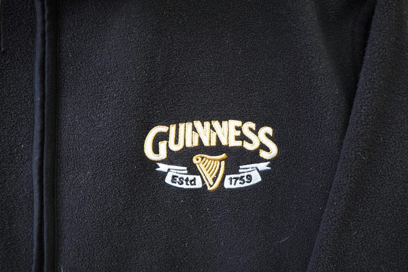 May include: Black fleece jacket with the Guinness logo embroidered on the chest. The logo features the Guinness name in gold lettering, a gold harp, and the text "Est. 1759".