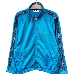 May include: A turquoise Fila track jacket with a full-length zipper and a stand-up collar. The jacket features the Fila logo on the chest and repeated on the sleeves. The sleeves have a black band with the Fila logo in multiple colors.