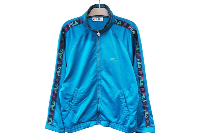 May include: A turquoise Fila track jacket with a full-length zipper and a stand-up collar. The jacket features the Fila logo on the chest and repeated on the sleeves. The sleeves have a black band with the Fila logo in multiple colors.