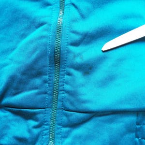 May include: Close-up of a teal-colored fabric with a zipper running vertically down the center. The fabric has a slight texture and appears to be part of a garment. A small wooden stick is in the upper right corner.