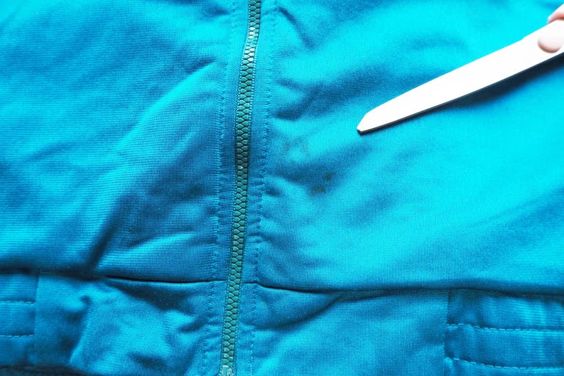 May include: Close-up of a teal-colored fabric with a zipper running vertically down the center. The fabric has a slight texture and appears to be part of a garment. A small wooden stick is in the upper right corner.