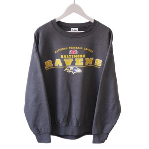 Może przedstawiać: Ciemnoszara bluza Lee z logo Baltimore Ravens. Napis "National Football League" i "Baltimore Ravens" jest w kolorze złotym i białym. Logo Ravens to fioletowy kruk. Bluza wisi na białym wieszaku.