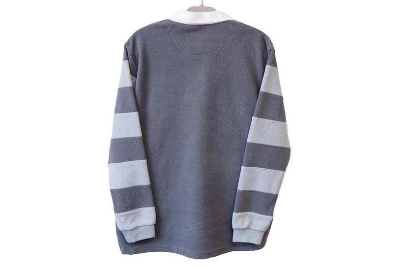 May include: A gray long-sleeved shirt with white and gray striped sleeves, a white collar, and a small patch on the back. The shirt is hanging on a white hanger against a white background.