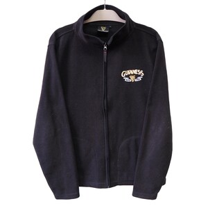 May include: Black fleece zip-up jacket with the Guinness logo embroidered on the chest. The logo features the Guinness name in white and gold, with a harp symbol. The jacket has a full zipper and two pockets.
