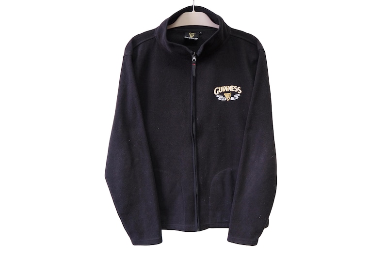 May include: Black fleece zip-up jacket with the Guinness logo embroidered on the chest. The logo features the Guinness name in white and gold, with a harp symbol. The jacket has a full zipper and two pockets.