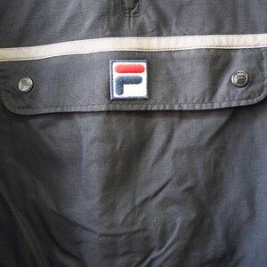 Op de afbeelding: Close-up van een donker marineblauwe jas met een zak en twee zilveren knopen. De zak heeft een rechthoekige patch met het Fila-logo in rood, wit en blauw. Een lichtbeige streep loopt over de borst.